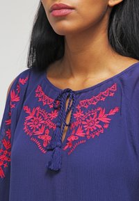 Vrouw in een blauwe blouse met rode bloemenborduursels en een franjes-sluiting bij de halslijn, zichtbaar zijn nek en bovenste deel van de borst.