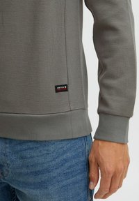 Siv sweatshirt od waffle pletenine s rebrastim rubom i manžetama, s crnom etiketom s crvenim detaljima na donjoj strani, u paru s plavim trapericama.