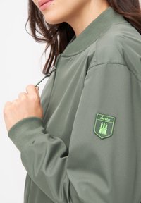 Grön bomberjacka med en slät textur, ribbade ärmslut och en framträdande grön badge med ett slottsdesign på vänster ärm.