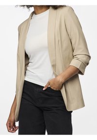 Beige blejzr s uvolněným střihem, ohrnutými rukávy a otevřeným předním designem, kombinovaný s bílým tričkem a černými kalhotami. Hladká textura.