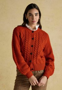 Adīta oranža džemperis ar kabeli modeļiem, ribotām piedurknēm un priekšējiem pogām. Valkāts pāri baltai bārkstainai krekla ar struktūru.