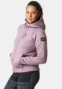 Lila zip-up hoodie met een hoge kraag, voorzakken en een zwart logo patch op de linkermouw. Gemaakt van zacht, rekbaar materiaal.