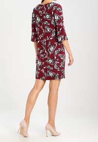 Robe rouge avec imprimé floral noir, manches trois-quarts, forme légèrement évasée, portée avec des talons hauts rose clair. Tissu lisse et léger.