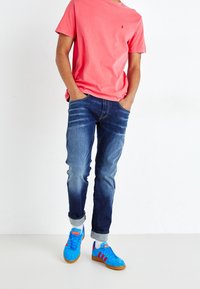 Rosa bomullströja, blå denimjeans med blekt framsida, blå mockaskor med röda ränder, allt på en enfärgad bakgrund.