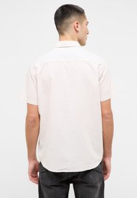 Koszula z krótkim rękawem w kolorze beżowym, zapinana na guziki, z klasycznym kołnierzykiem, miękką teksturą i zaokrąglonym dołem. Widok z tyłu ukazuje gładki, minimalistyczny design.