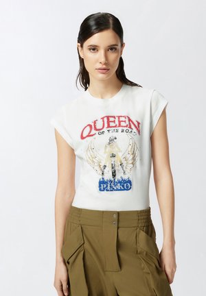 Maglietta grafica bianca con stampa "QUEEN OF THE ROAD" in rosso, caratterizzata da un design con ali; stile aderente, indossata con pantaloni verde oliva.