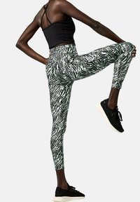 Top atletico nero abbinato a leggings a vita alta con un motivo astratto verde e nero. La modella indossa sneakers nere.
