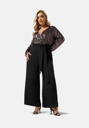 Femme portant un haut noir à sequins à manches longues, associé à un pantalon noir taille haute à jambes larges et des sandales noires à talons hauts.