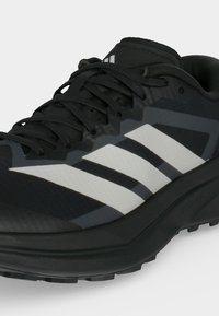 Μαύρο αθλητικό παπούτσι με λευκό σχέδιο τριών ρίγων, μαύρα κορδόνια και υφή στην σόλα, εμφανιζόμενο από μπροστινή πλευρική γωνία.