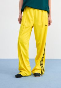 Yellow polyester track pants with black side stripes, featuring a loose fit and two front pockets. The waistband is elasticized.