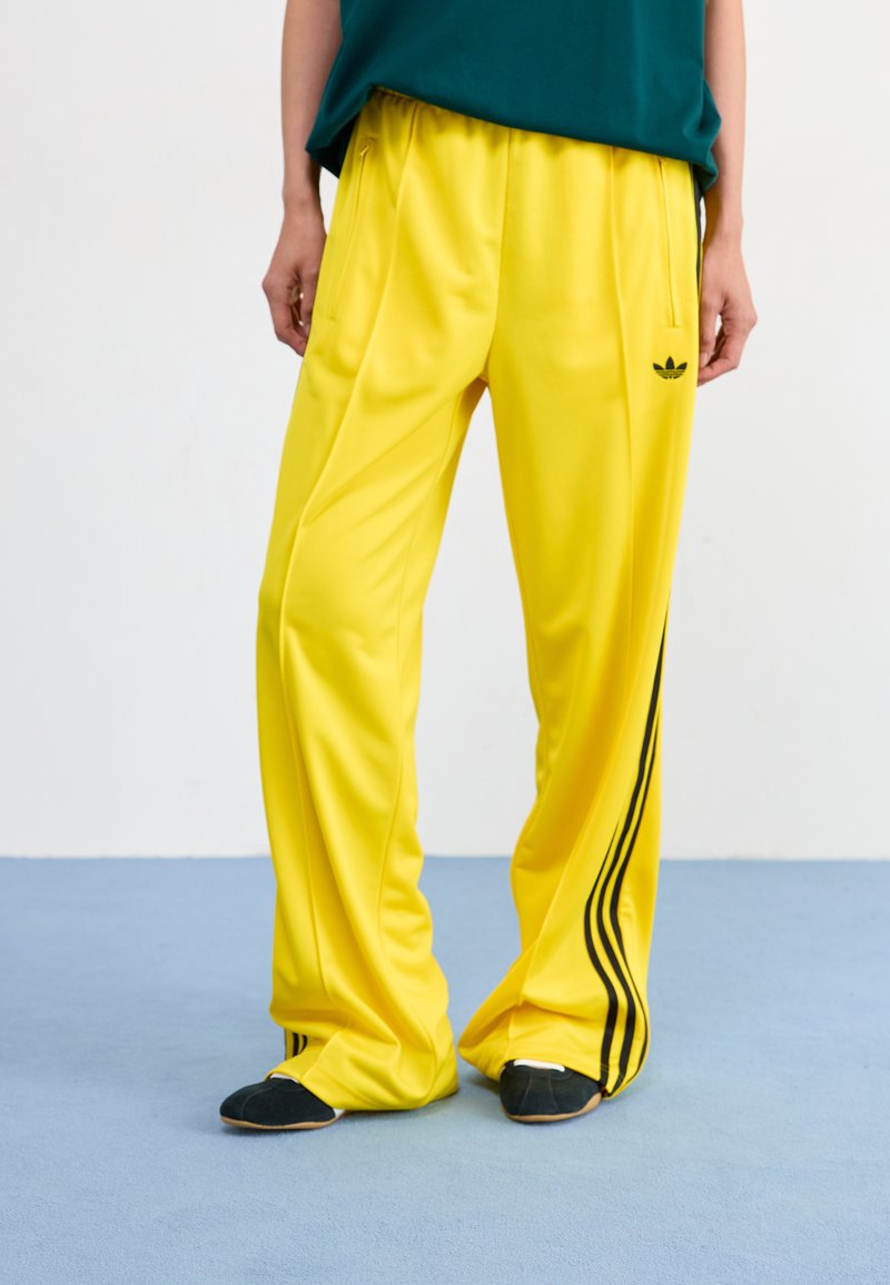 Yellow polyester track pants with black side stripes, featuring a loose fit and two front pockets. The waistband is elasticized.
