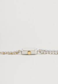 Ceas argintiu cu o carcasă rectangulară, lanțuri de legătură din aur și argint, și pietre decorative transparente de-a lungul curelei. Buton auriu îndrăzneț pe lateral.