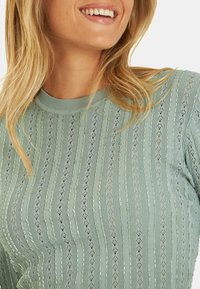 Hellgrüner Strickoberteil mit vertikalen Streifen und rautenförmigen Mustern. Rundhalsausschnitt, halbdurchsichtige Textur und gerippte Bündchen.