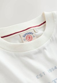 Biela bavlnená tričko s kruhovým výstrihom, rebrovaným golierom s červeným akcentom a modrou tlačenou nášivkou vo vnútri výstrihu.