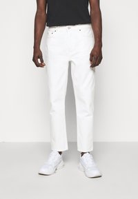 Białe dżinsy o prostych nogawkach, wykonane z gładkiego denimu, z klasycznym zapięciem na guzik i przednimi kieszeniami. Noszone z białymi trampkami.