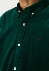 Zielona koszula z sztruksu zapinana na guziki, z teksturowanym wykończeniem, białymi guzikami oraz małym czarnym logo na piersi.