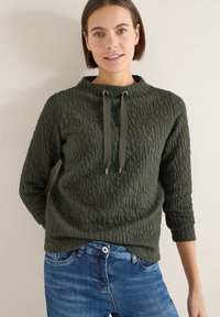 Mujer vistiendo una blusa de manga larga texturizada en verde oscuro con cuello ajustable mediante cordón y jeans azules, de pie contra una pared de color beige claro.