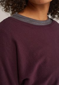 Bordeauxfarbener Pullover mit gerippter Struktur, der einen weiten Ausschnitt hat. Ein graues Unterziehoberteil ist sichtbar, was einen Kontrast zur Farbe bietet.