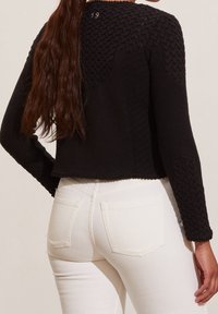 Černý pletený svetr s texturovanými rukávy a krátkým střihem, kombinovaný s bílými džíny. Má složité vzory na ramenou a manžetách.
