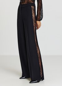 Pantaloni neri a gamba larga con pannelli laterali in pizzo, vita liscia e texture elegante. Presentano tacchi neri lucidi che completano il look.