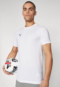 Biała koszulka sportowa z krótkim rękawem, okrągłym dekoltem i szarymi akcentami. Model trzyma czarno-białą piłkę nożną z czerwonymi detalami.