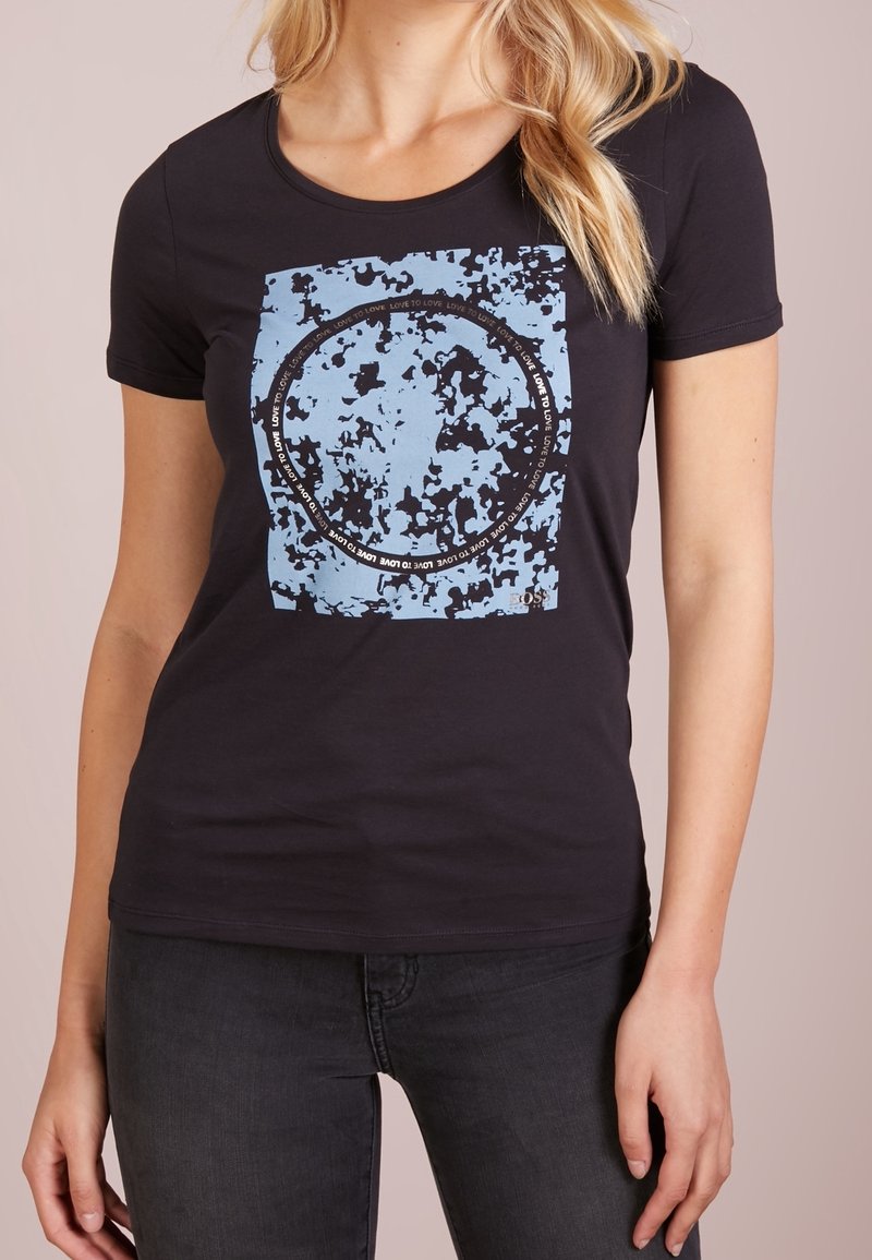 Camiseta de algodón negra con un diseño gráfico de cuadrados azules que presenta un patrón de texto circular y acentos desgastados. Manga corta, corte ajustado.