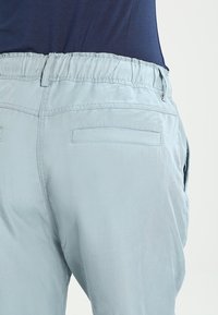 Světle modré kalhoty s elastickým pasem, dvěma zadními kapsami a hladkým povrchem. Tkanina se zdá být lehká a mírně lesklá.