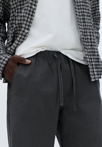 Grå bukser med elastisk talje og snøre; sat sammen med en hvid t-shirt og en sort-hvidternet skjorte, hvilket viser et afslappet, lagdelt look.