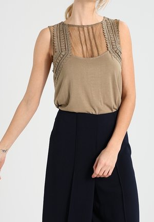 Vrouw draagt een mouwloos beige topje met kant en doorschijnende gestreepte details, gecombineerd met een zwarte broek met hoge taille, tegen een effen achtergrond.