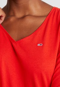 Rode katoenen V-hals top met een slanke pasvorm en een klein geborduurd logo aan de linkerkant. Gladde textuur met subtiele stiksel details.