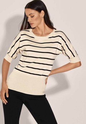 Frau trägt einen cremefarbenen Pullover mit schwarzen horizontalen Streifen und schwarze Hose, posiert mit einer Hand auf der Hüfte und schaut seitlich nach unten.