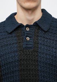 Sötétkék és fekete kötött pólóing, texturált mintával, gallérral és három gombos záródással a nyaknál.