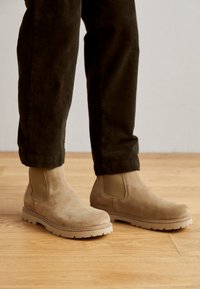 Botas Chelsea de ante beige con paneles laterales elásticos, suelas de goma y puntera redondeada. Combinadas con pantalones de pana oscuros sobre una superficie de madera.