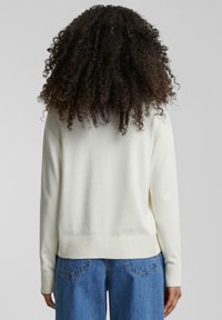 Pull léger beige avec un ourlet et des poignets côtelés, présentant un design simple. Le dos présente une texture douce et une coupe décontractée associée à un jean bleu.