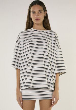 Kvinde med langt brunt hår iført en stor hvid skjorte og et kort nederdel, begge med tynde sorte horisontale striber, stående mod en ensfarvet baggrund.