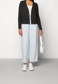 Cardigan grigio a coste, top corto bianco, jeans a gamba larga azzurri chiari e sneakers bianche, con una piccola borsa bianca in mano.