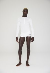 Hvid langærmet top med knaplukning, tætsiddende design og dekorativ syning, parret med tætsiddende hvide trusser. Model står barfodet foran en neutral baggrund.