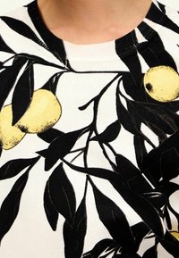 Zbliżenie na biały materiał z czarnymi liściastymi gałęziami i nadrukowanymi żółtymi owocami przypominającymi cytryny na dekolcie odzieży.