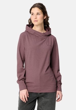 Frau mit dunklen Haaren, die einen weinroten Kapuzenpullover und dunkle Hosen trägt, steht vor einem schlichten hellen Hintergrund.