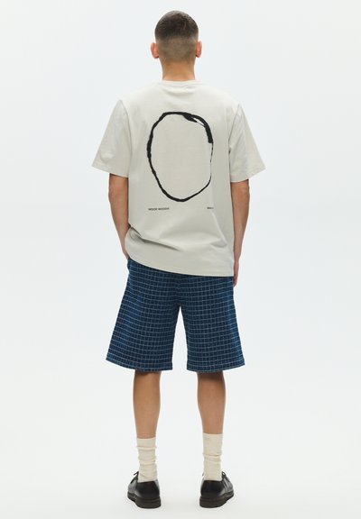 Man wearing white t-shirt with black oval design, blue checkered knee-length shorts, cream socks, and black shoes, standing facing away.