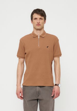 Férfi barna rövid ujjú, félcipzáras galléros pólót és szürke nadrágot visel, egyszínű világos háttér előtt áll.