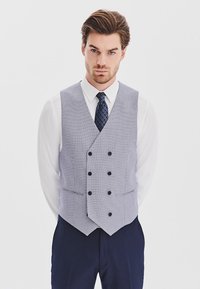 Szary i biały kamizelka w kratę z dekoltem w serek, podwójnym zapięciem z przodu i czarnymi guzikami. Nosi się ją na białej koszuli i granatowym krawacie.