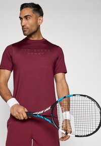 Bordo sportinė marškinėlė su apvalia iškirpte ir iškaltu logotipu, derinama su riešo raišteliais. Modelis laiko tenisą raketę su juodu ir mėlynu rėmu.