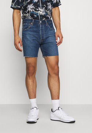 Denim shorts i mellemblå med en rå kant, standardlommer og lynlås, parret med en kortærmet skjorte med tropisk print.