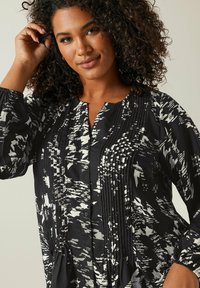 Blusa a motivi in bianco e nero con placca frontale, dettagli plissettati, polsini elasticizzati e vestibilità comoda. Tessuto dalla texture liscia.