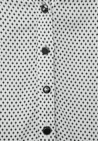 Chemise noire et blanche à boutons, présentant un motif à pois, un tissu texturé, six boutons noirs sur le devant, et un col classique.