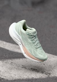 Ανοιχτό πράσινο αθλητικό παπούτσι με λευκή και ροδακινί σόλα, στον αέρα πάνω από επιφάνεια από σκυρόδεμα, με μικρά θραύσματα να αιωρούνται γύρω του.