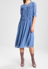 Robe bleue, longueur genoux, avec un buste froncé, des manches longues et une jupe fluide. Portée avec des bottines mauves. Texture lisse.