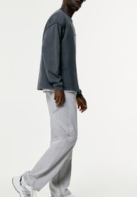 Felpa grigia a maniche lunghe, indossata con jeans grigi chiari. Sneakers nere con dettagli bianchi. Il modello è in piedi su uno sfondo neutro.