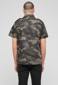 Korte mouwen camouflage shirt in donkere groene, grijze en beige tinten, met een standaard kraag en schouderepauletten voor detailing.